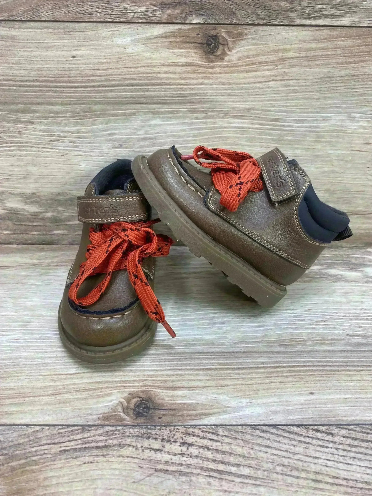 Front and side view of Carter’s Norman ankle boots in brown with orange laces, velcro strap, and treaded sole on wood