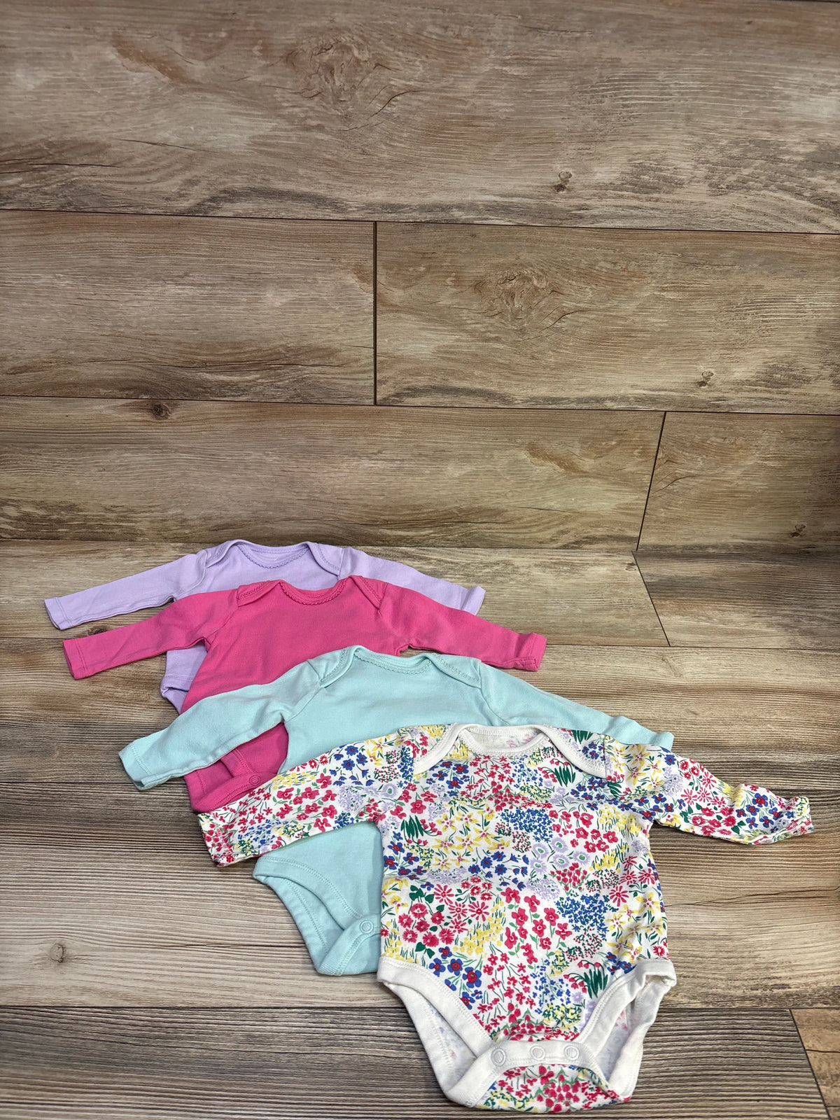 Top view of Wonder Nation 4-pack bodysuit set showcasing pink, lavender, aqua, and floral-print bodysuits on wood