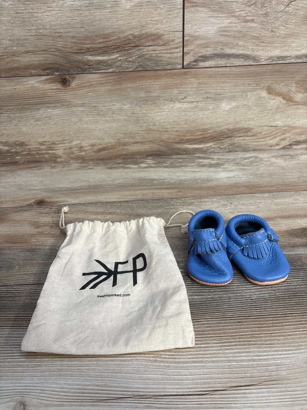 Blue baby moccasins next to Freshly Picked branded drawstring bag on wood floor