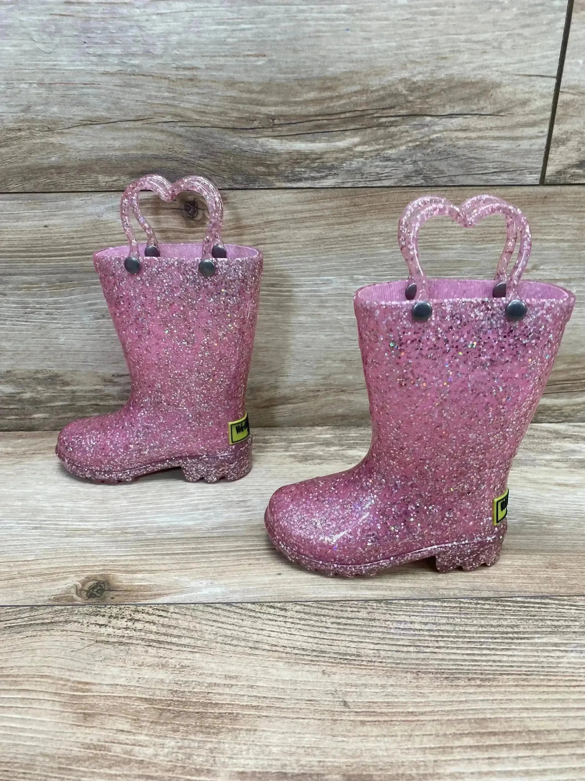 Side view of Western Chief Pink Glitter Light Up Rain Boots size 5c showcasing sparkly pink finish with heart pull handles on wood plank floor