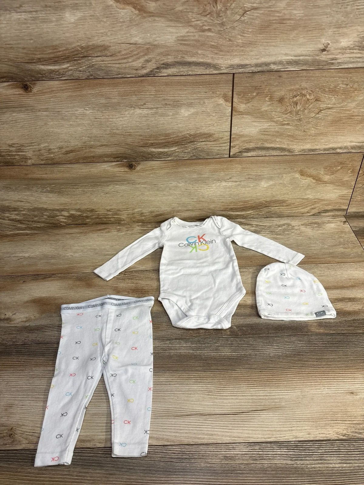 Calvin Klein baby outfit set with white onesie, pants, and hat, laid on wood floor