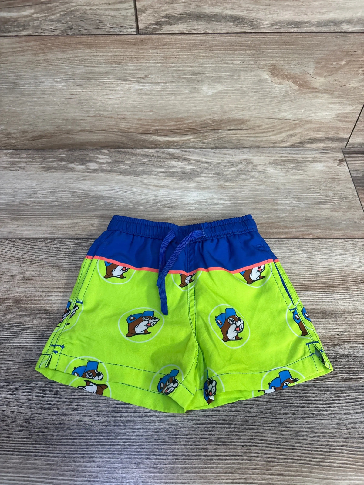 Front view of baby swim trunks, lime green with Buc-ee’s beaver logo, blue drawstring waistband on wood background