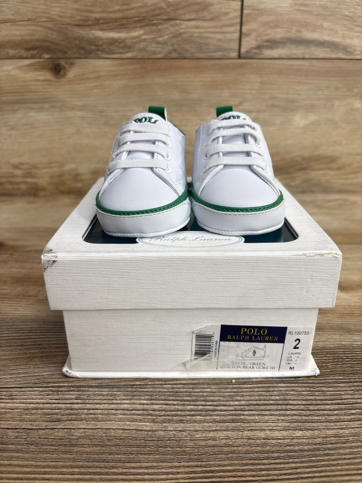 White and green Polo Ralph Lauren baby shoes on a white box with size 2 label, wooden background