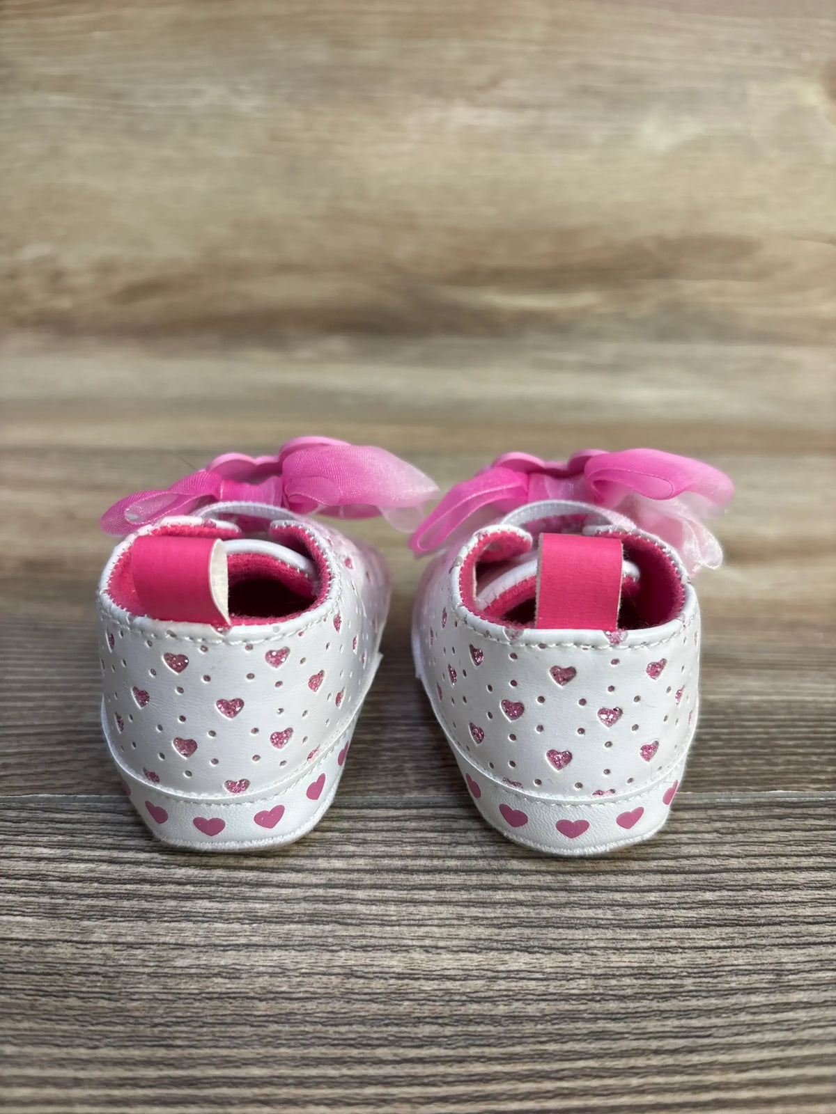 Rear view of Barbie Soft Sole Sneakers size 1C showcasing pink heart cutouts, glitter accents, and pink organza bows on a wood surface