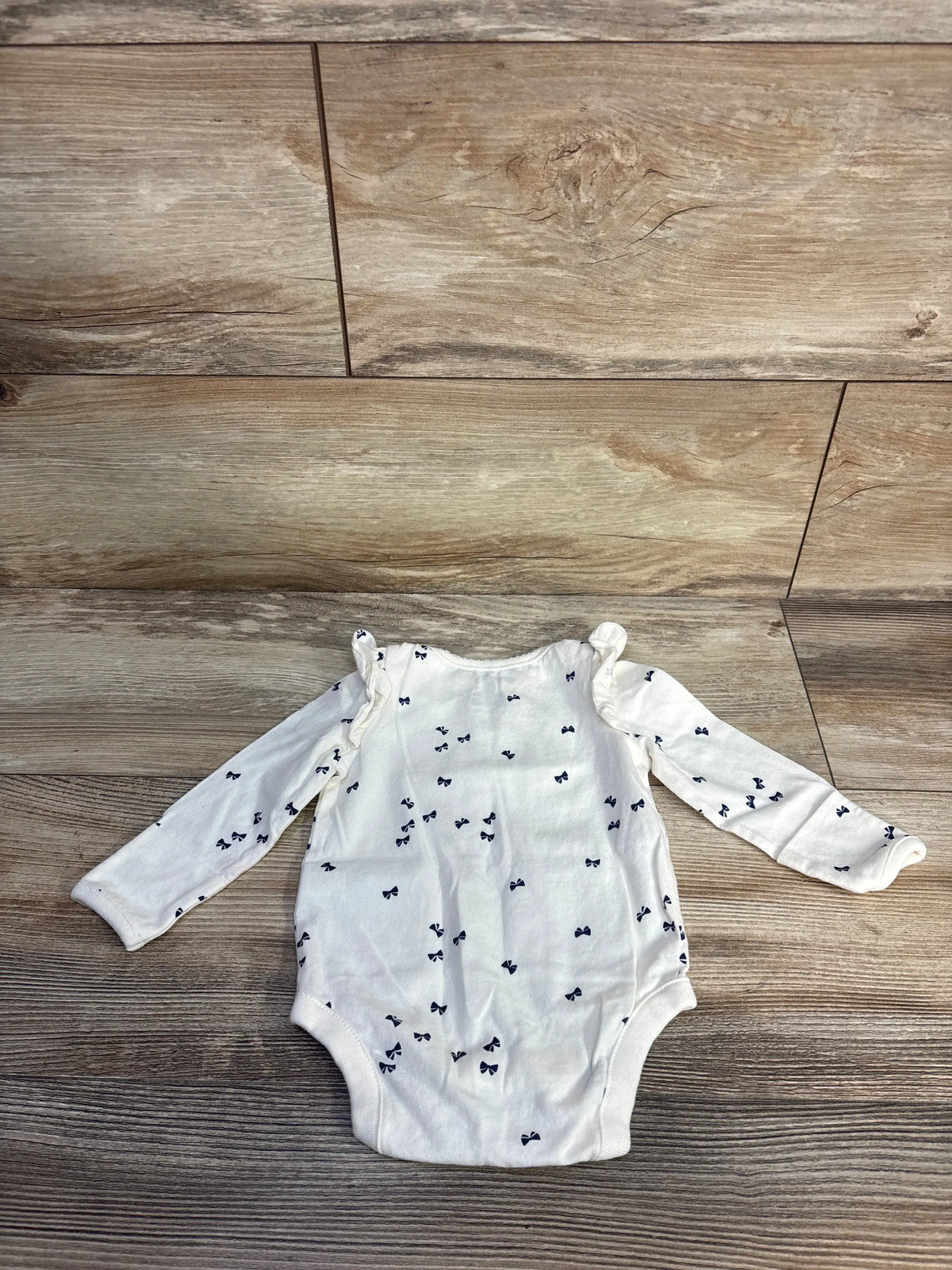 Front view of Baby Gap cream long sleeve bodysuit showcasing navy bow print and ruffled shoulders on a wood-grain floor