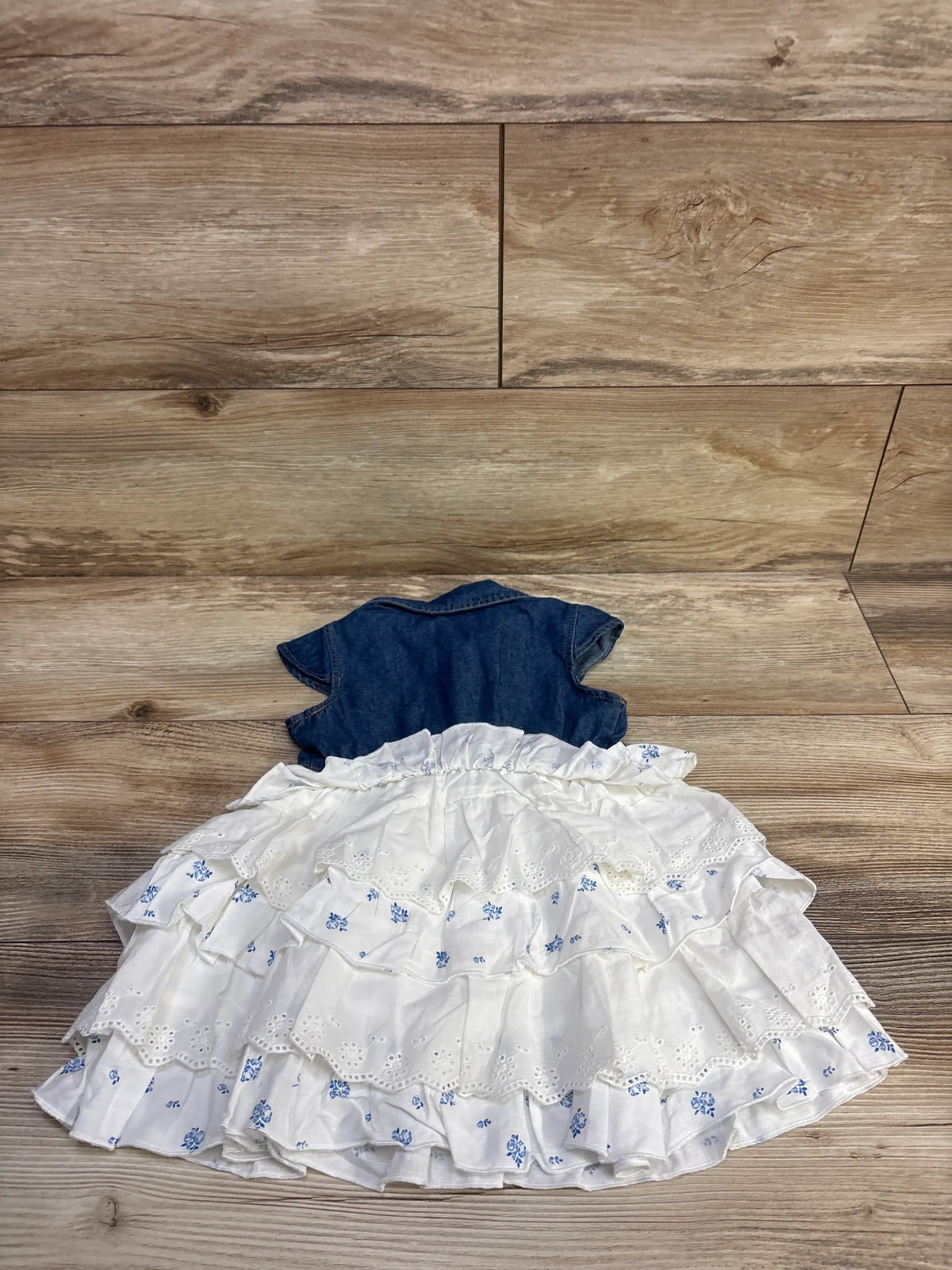 Flat lay of Lilt denim tiered skirt dress showcasing a blue denim bodice, white floral tiers, and eyelet trim on wood flooring