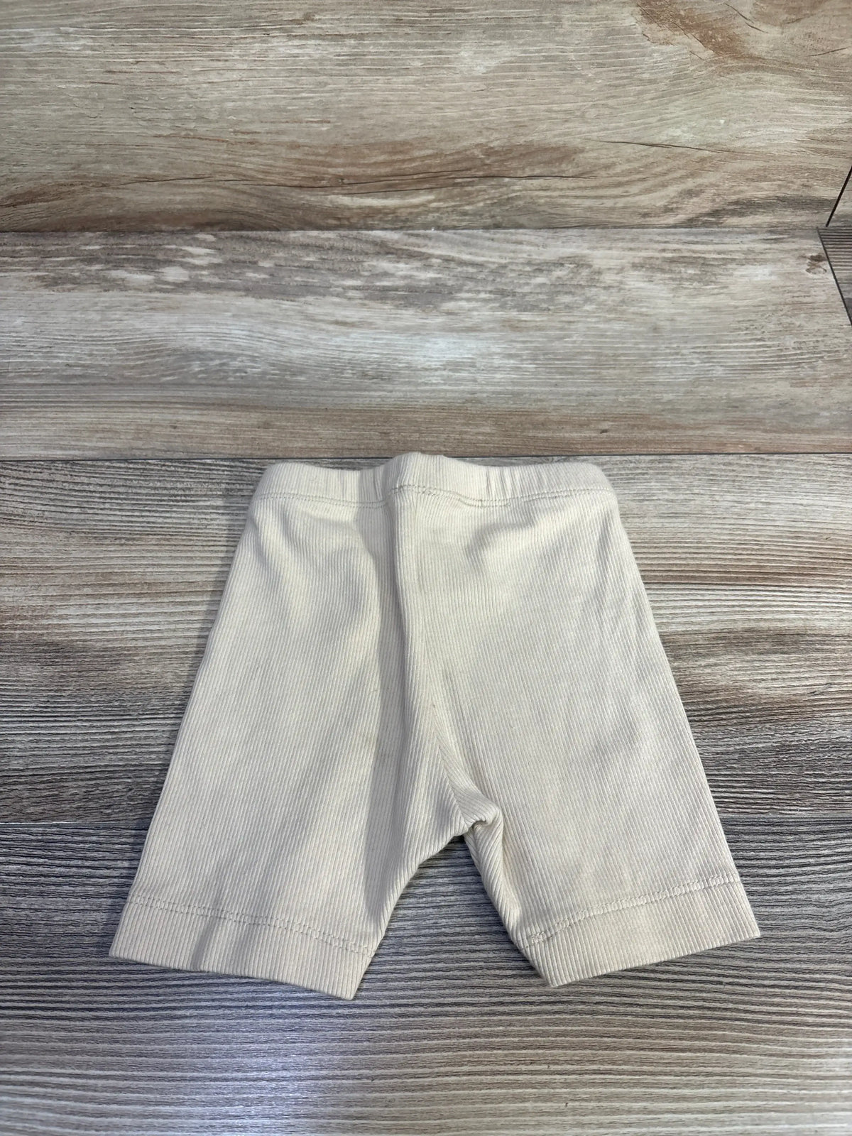 Flat lay of cream ribbed baby shorts (18-24 months) showing elastic waistband and knee-length legs on light wood background