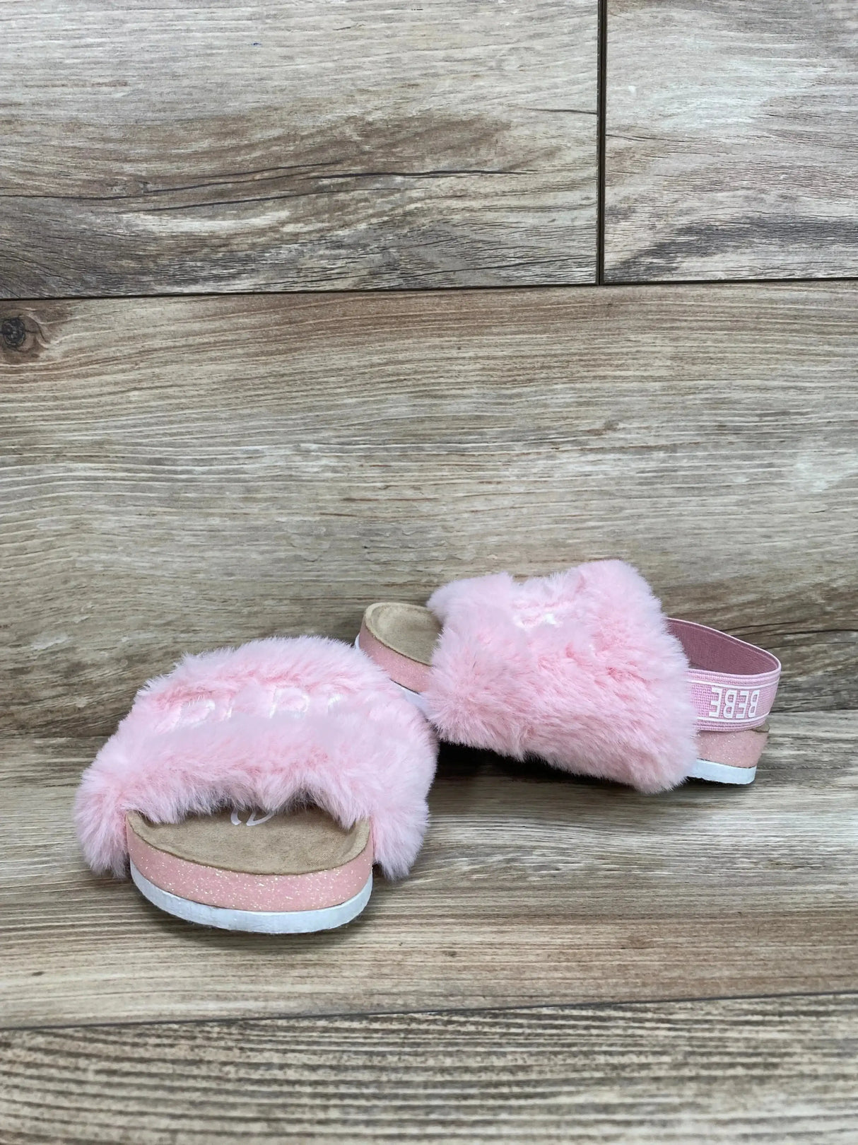 Front-angle of Bebe pink faux fur sandals (size 6C) showcasing fluffy pink upper, elastic branded heel strap, and glittery pink midsole on wood plank background