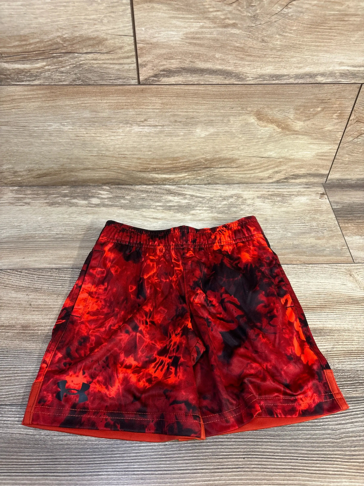 Flat lay of size 4T toddler red Under Armour athletic shorts with black flame-like print and elastic waistband on light wood floor