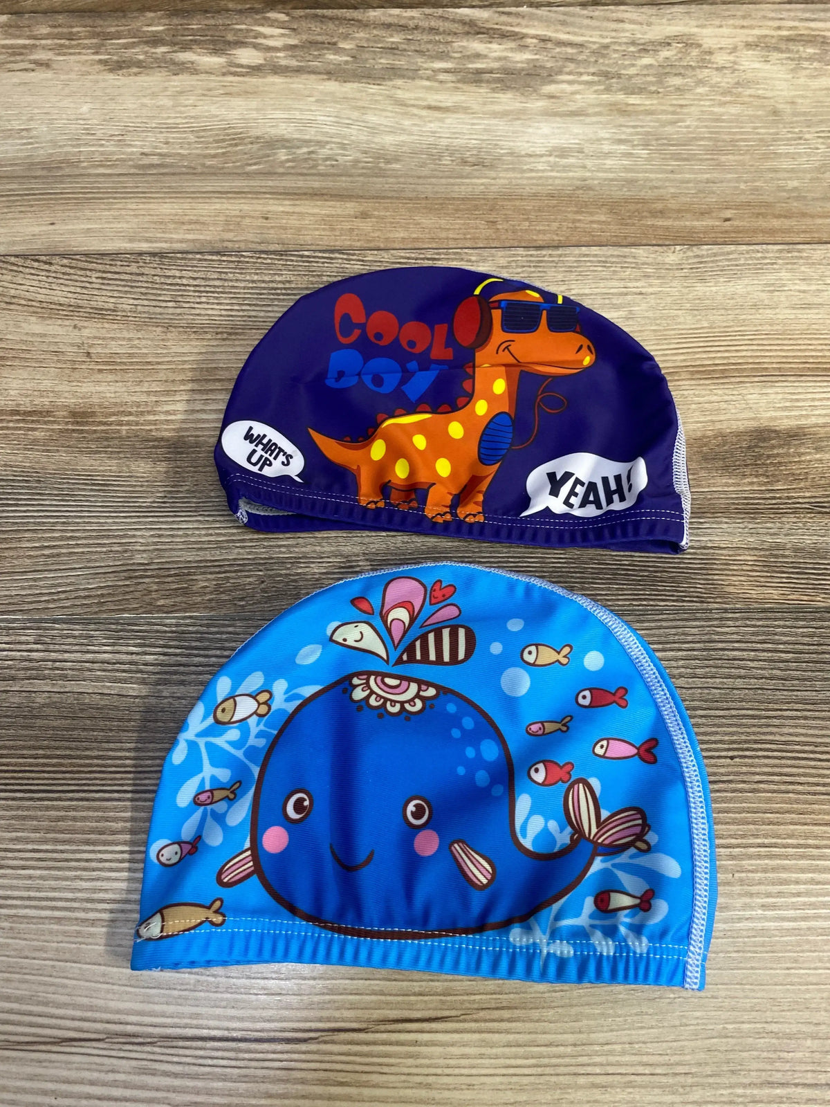 Top view of two children’s swim caps on wood: purple dinosaur cap labeled COOL BOY and blue whale cap