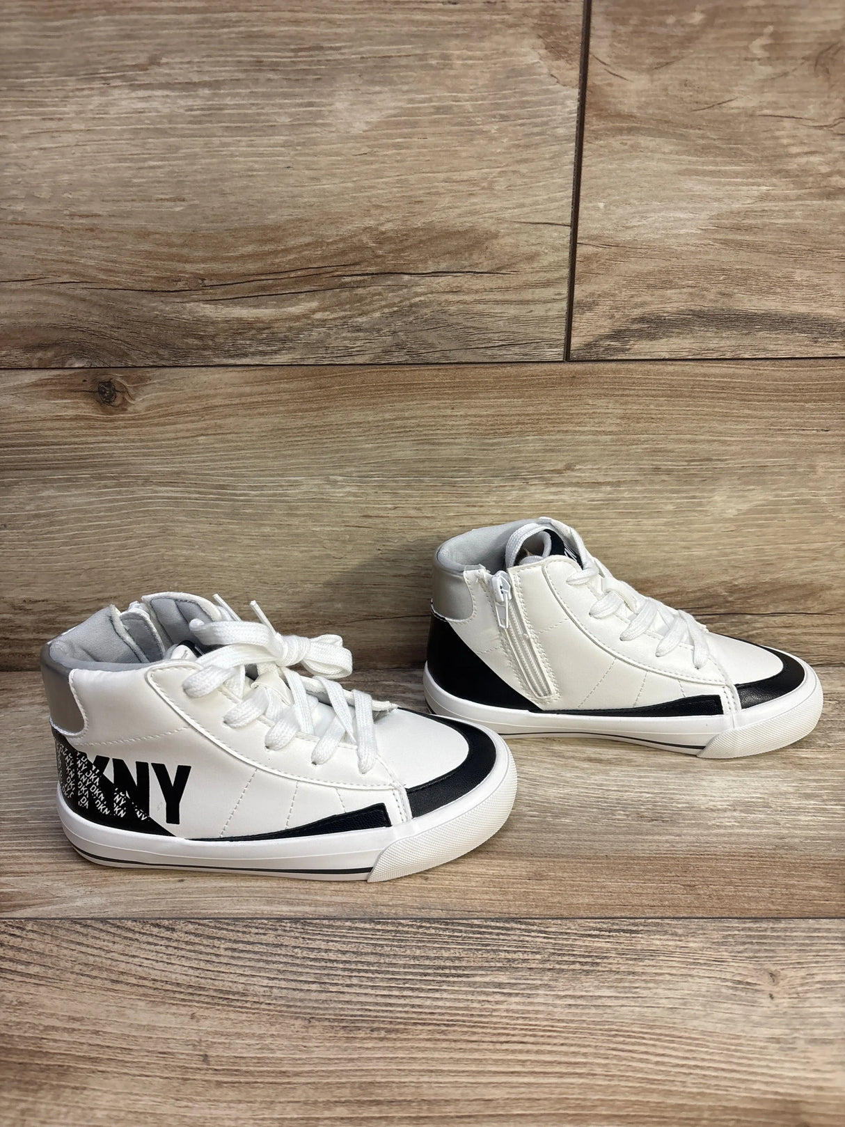 Side view of white DKNY Hannah Leena high-top girls sneakers with black trim, side zipper, laces, and DKNY logo on wood floor