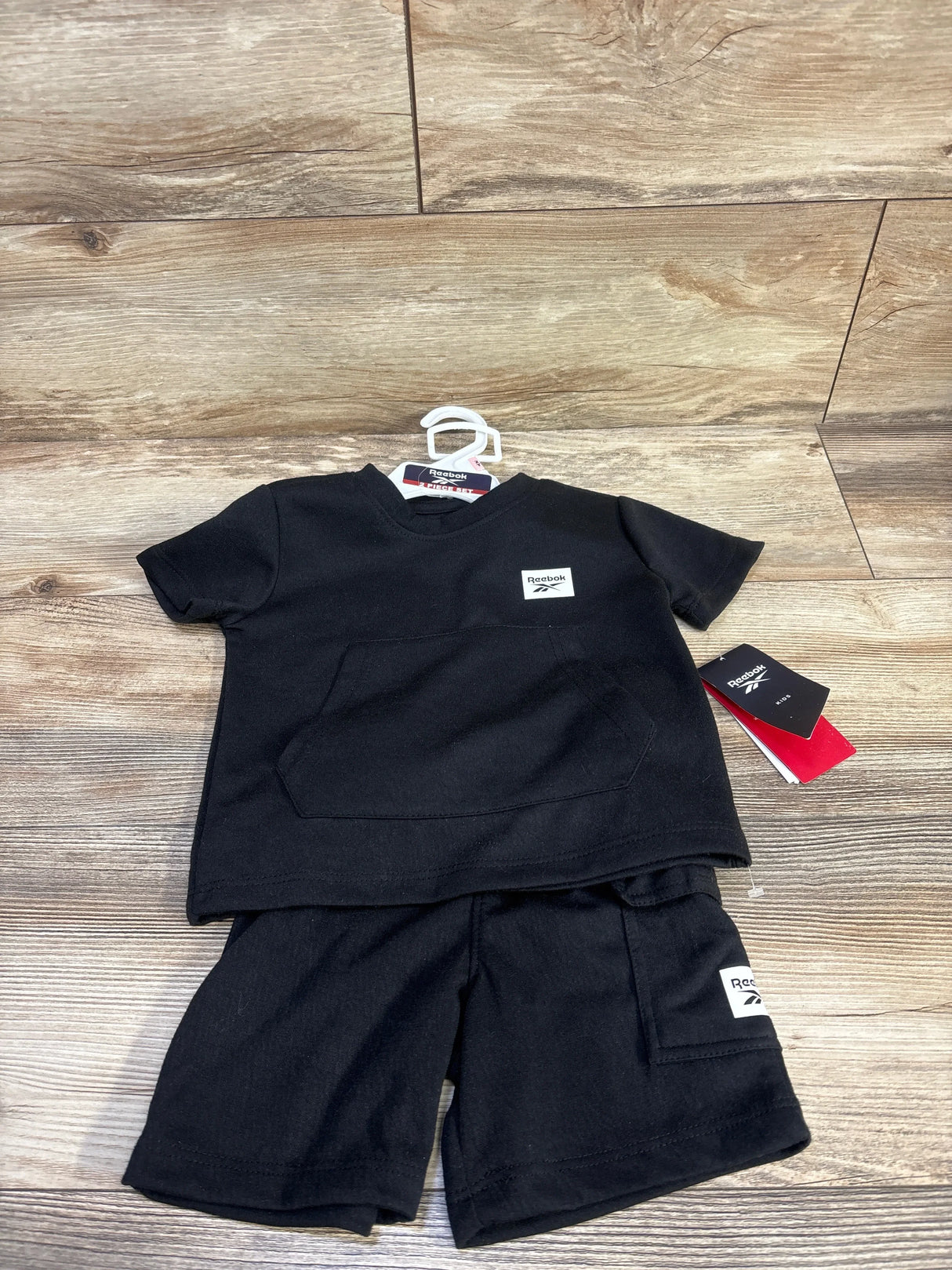 Flat-lay of black Reebok 2T shirt and shorts set showing front kangaroo pocket and Reebok logo patch on wood background