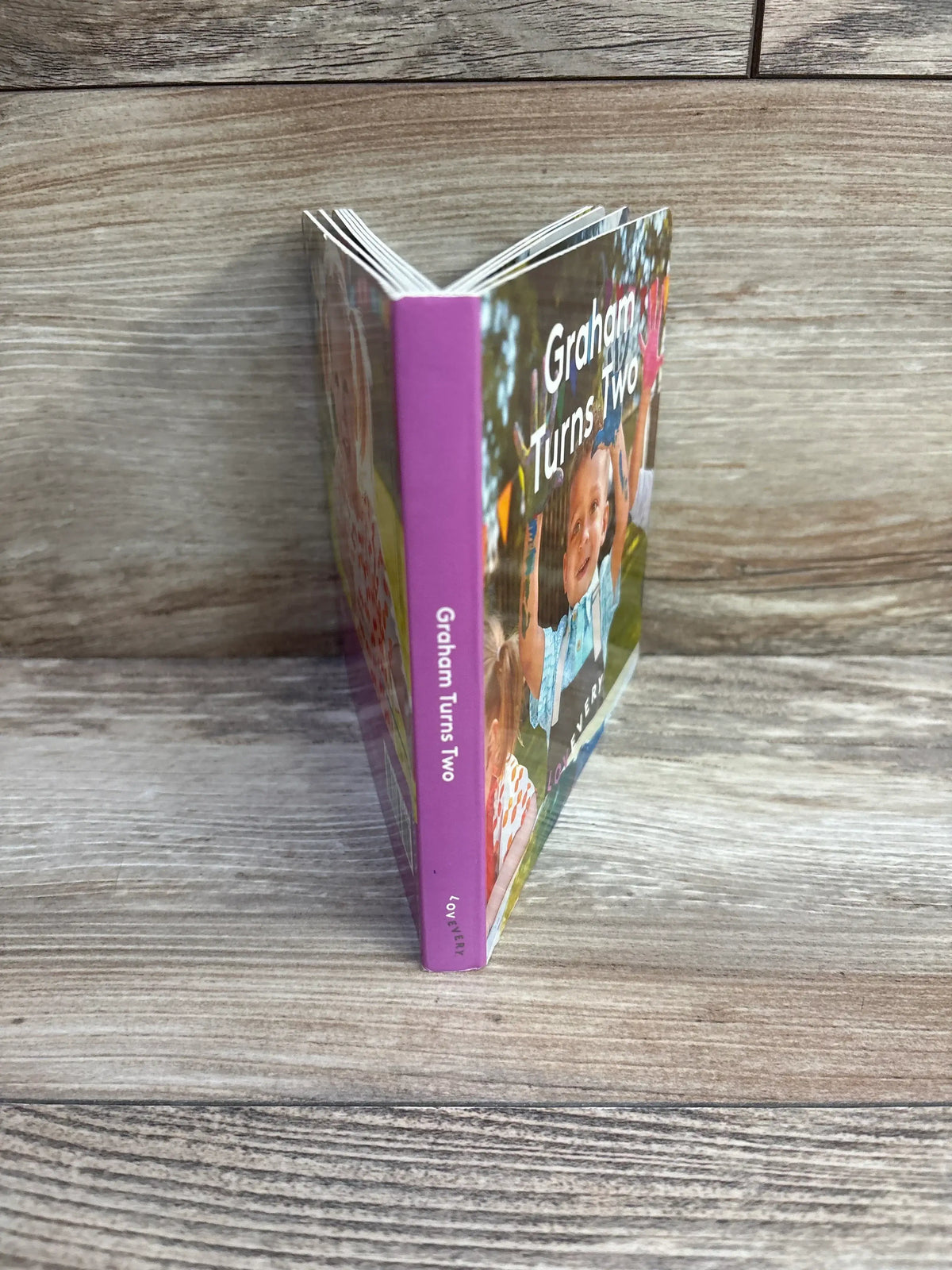 Angled front view of Lovevery Graham Turns Two board book with purple spine and cover photo of a toddler on wood background