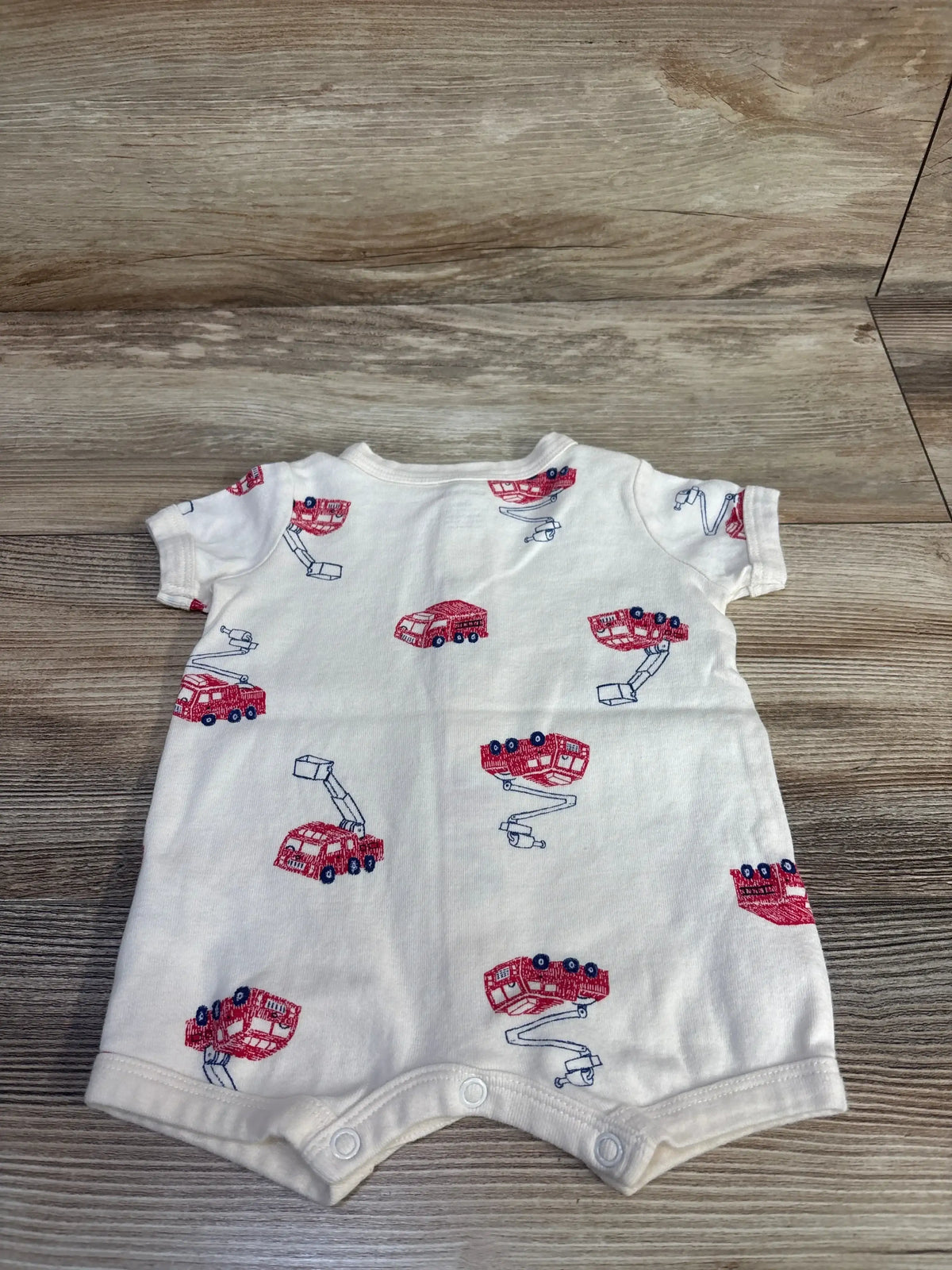 Flat lay of cream short-sleeve baby shortie romper with red fire truck print and front snaps on wood background
