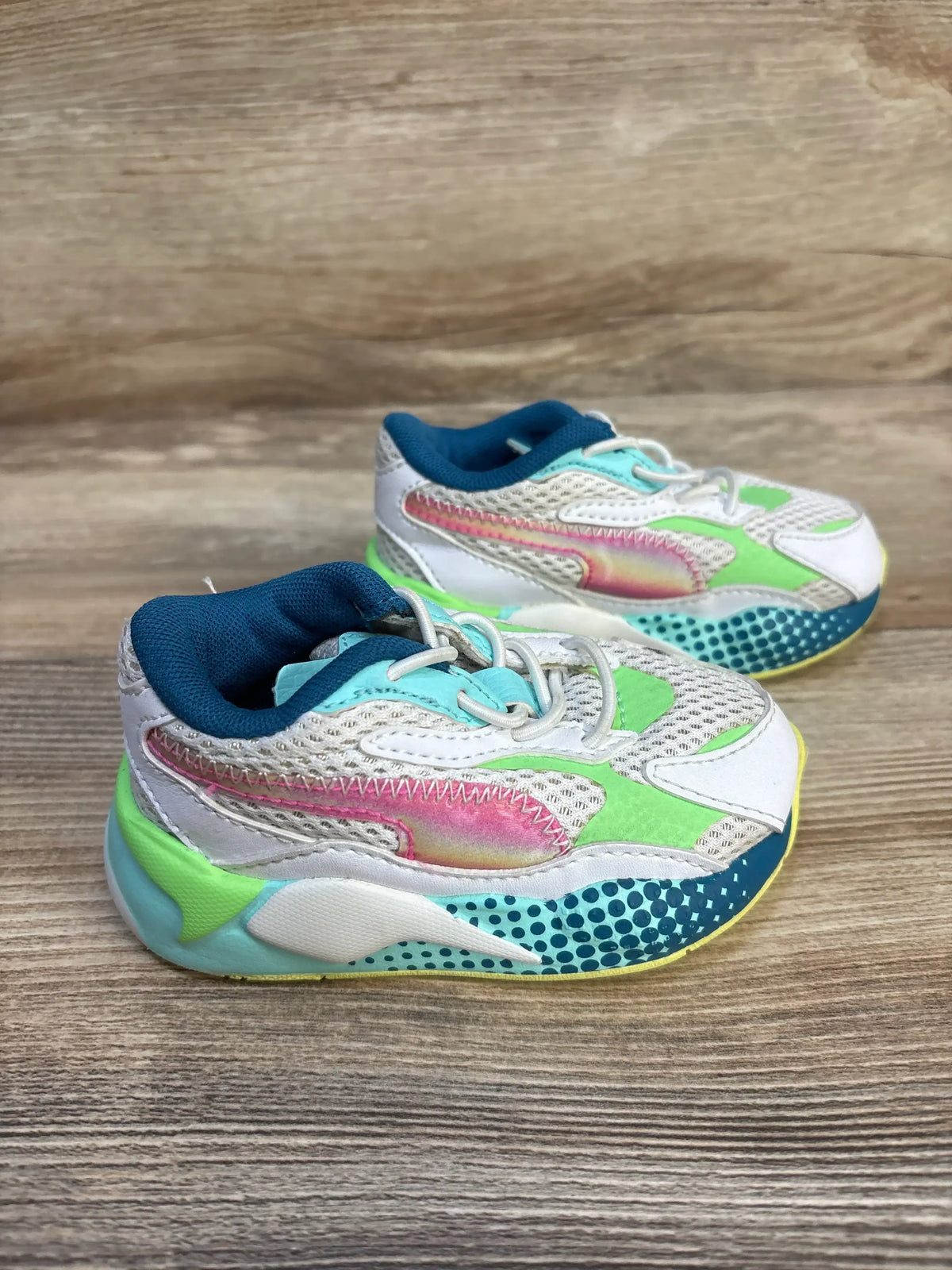 Three-quarter view of Puma RS-X3 toddler sneakers size 5C with white mesh, iridescent pink stripe and green-teal accents on wooden floor