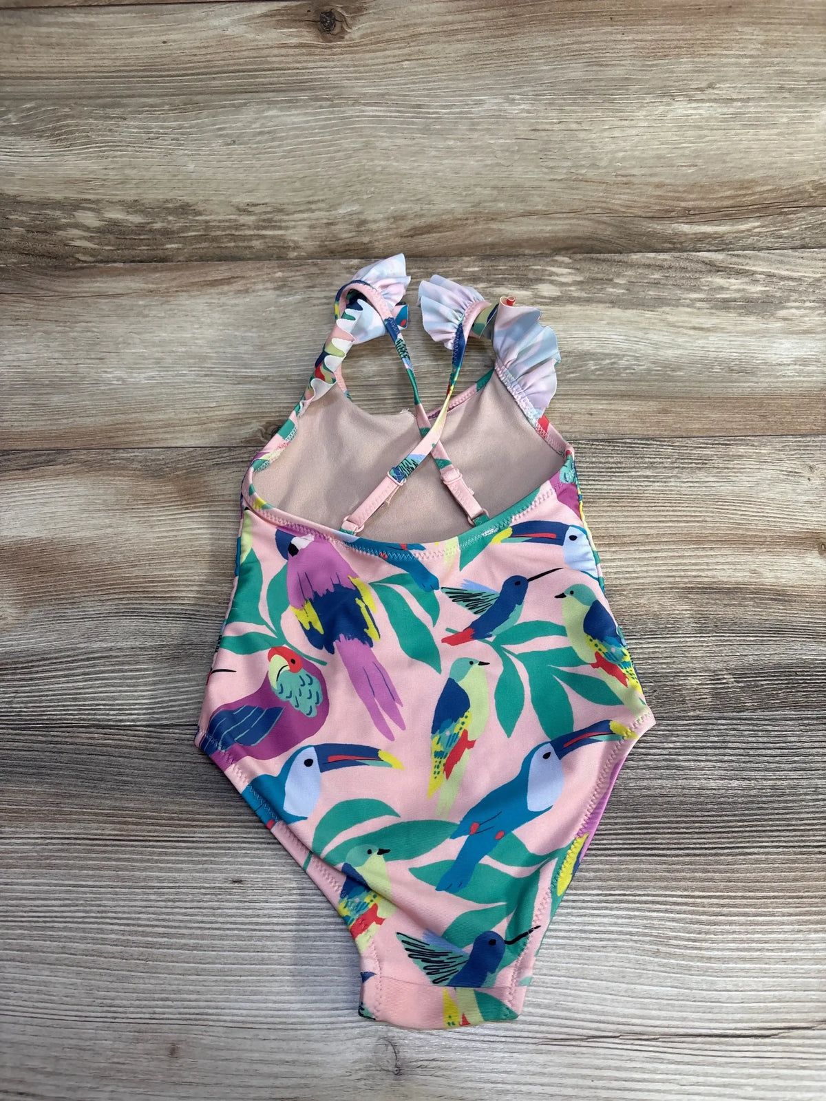 Flat lay of pink TEA one-piece swimsuit size 4T with ruffle straps and multicolored bird print on wood background