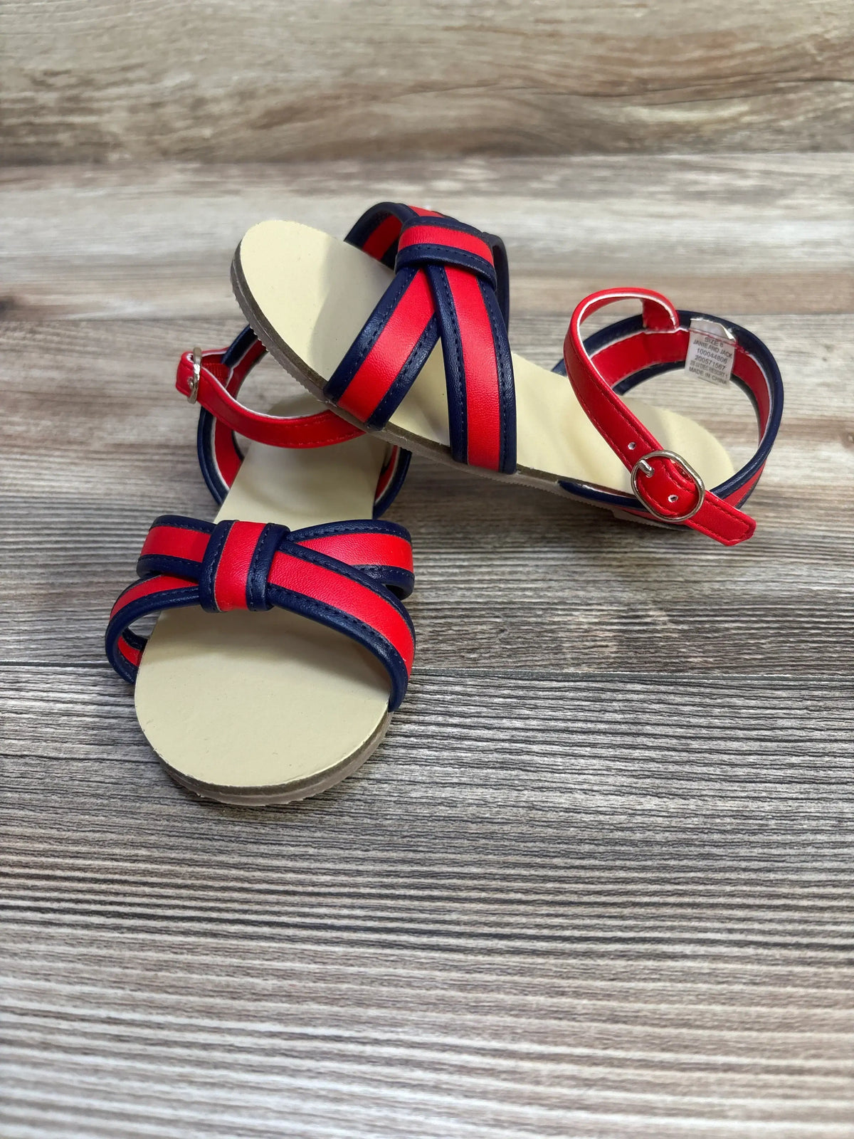 Front view of Janie and Jack toddler sandals with red and navy cross-straps, ankle buckle and beige sole on wood background