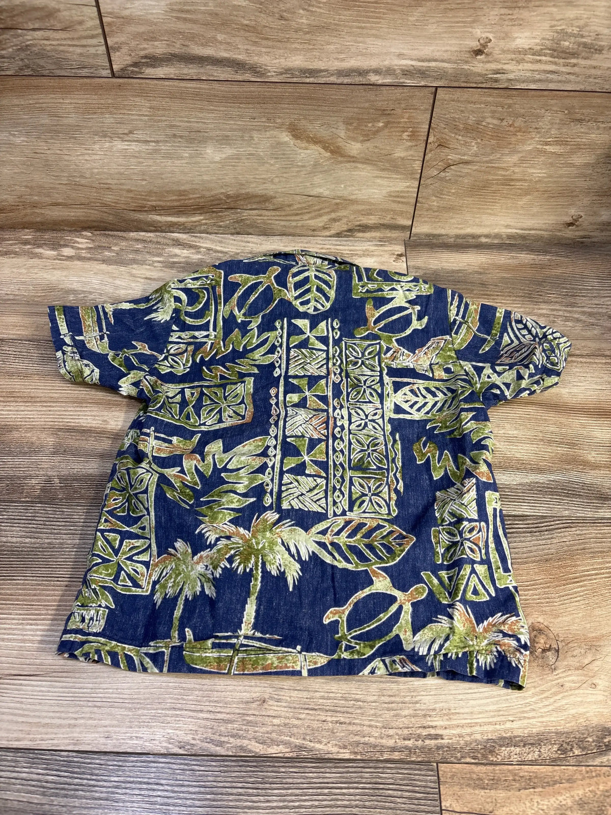 Back view of Bishop St. Hawaiian toddler short sleeve shirt showing green and beige tropical print on blue fabric on wood floor