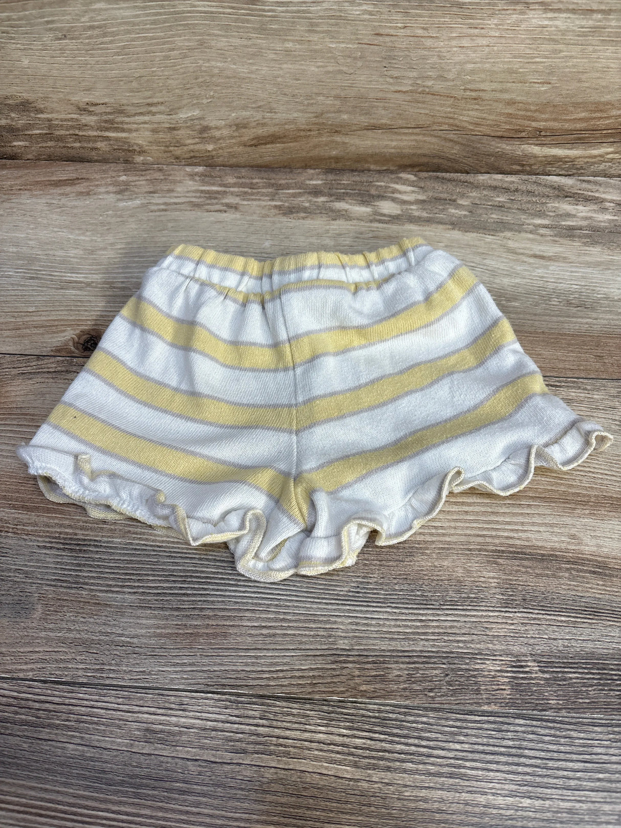 Flat lay of white shorts with pale yellow horizontal stripes, ruffled hem and elastic waistband, size 18 months, on wood background