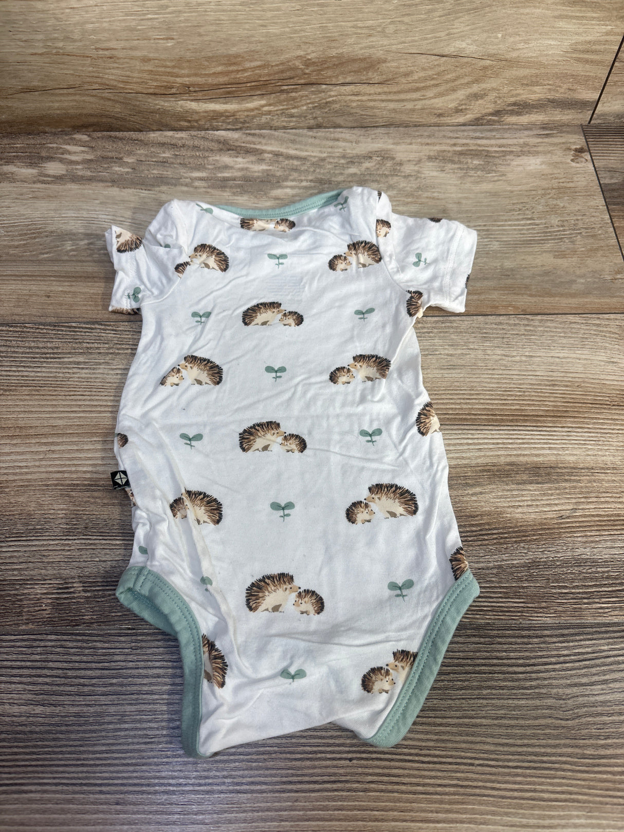 Top-down view of newborn white Kyte Baby short-sleeve bodysuit with hedgehog print and mint trim on wood background size 0-3m