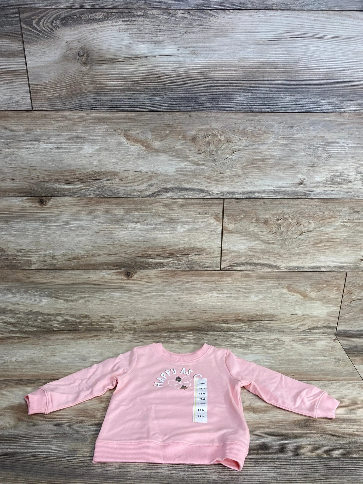 Flat lay of pink Cat and Jack toddler pullover sweater showing Happy As text, bee graphic and 12M size tag on wood background