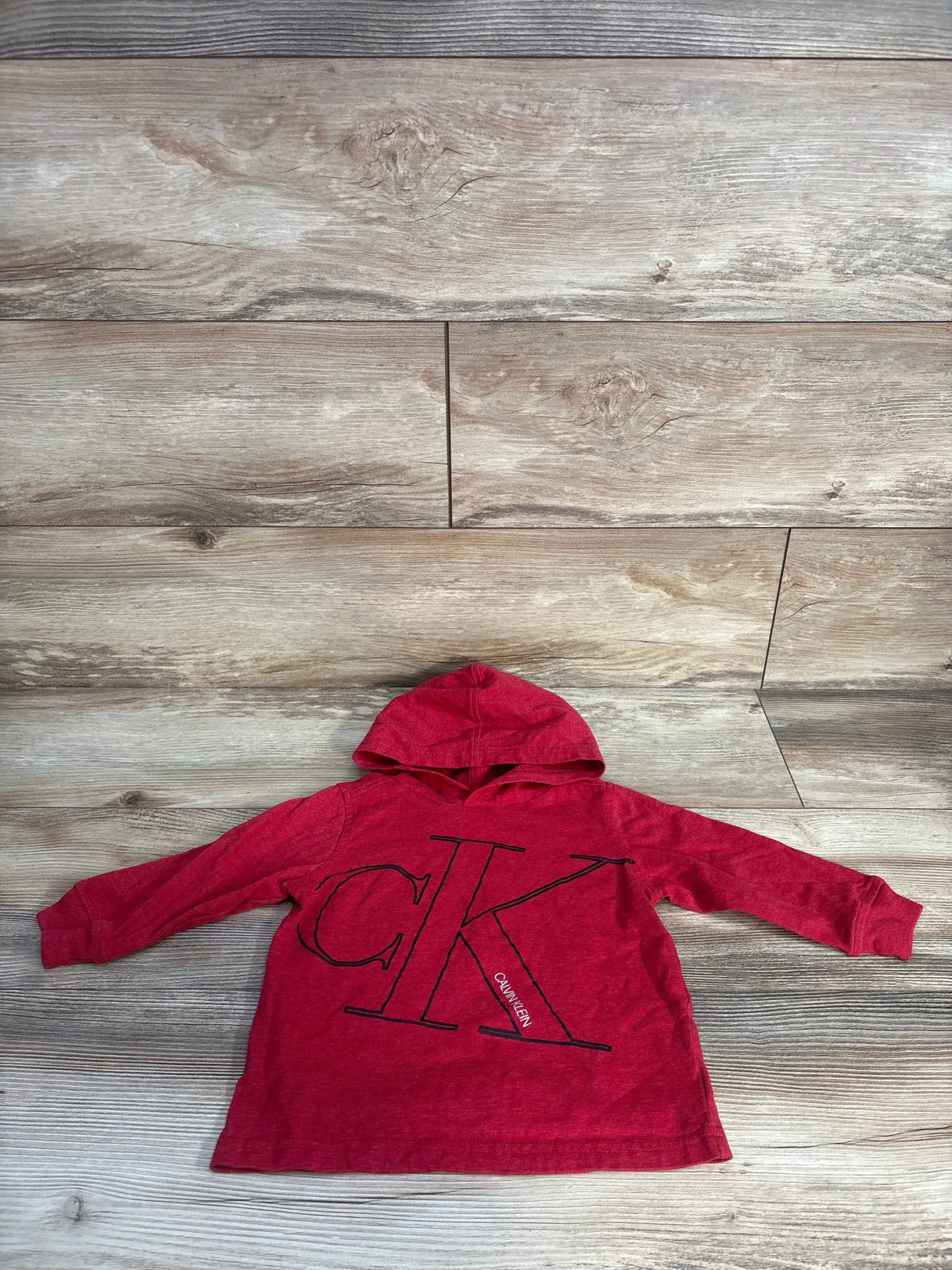 Flat lay of red Calvin Klein toddler hooded shirt size 2T showing large black CK logo on light wood background
