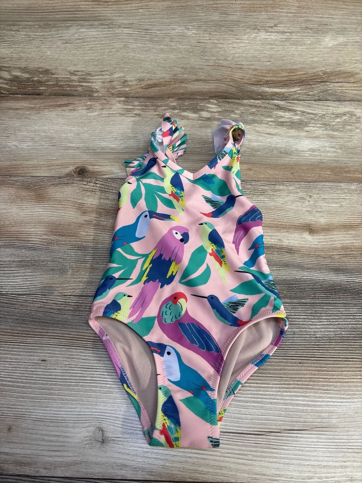 Flat lay of pink TEA girls one-piece swimsuit with colorful bird print and ruffle shoulder on wood background