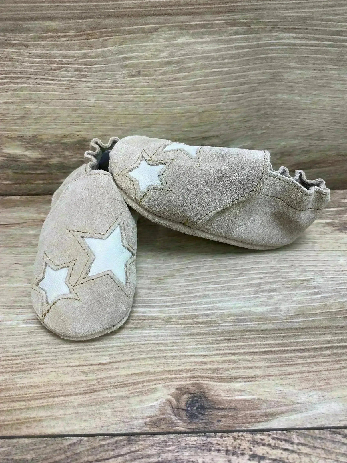 Top-front view of Minnetonka infant booties in tan suede showcasing white star appliqués and elasticized ankle on light wood background