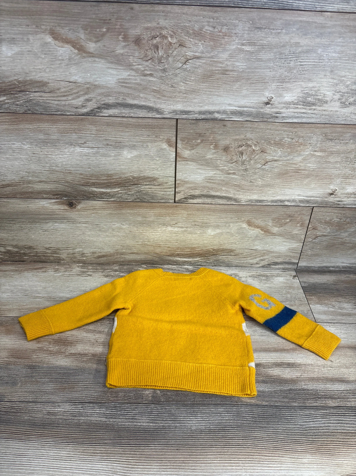 Yellow knit toddler sweater with blue stripe and letter G on sleeve, laid on wood floor