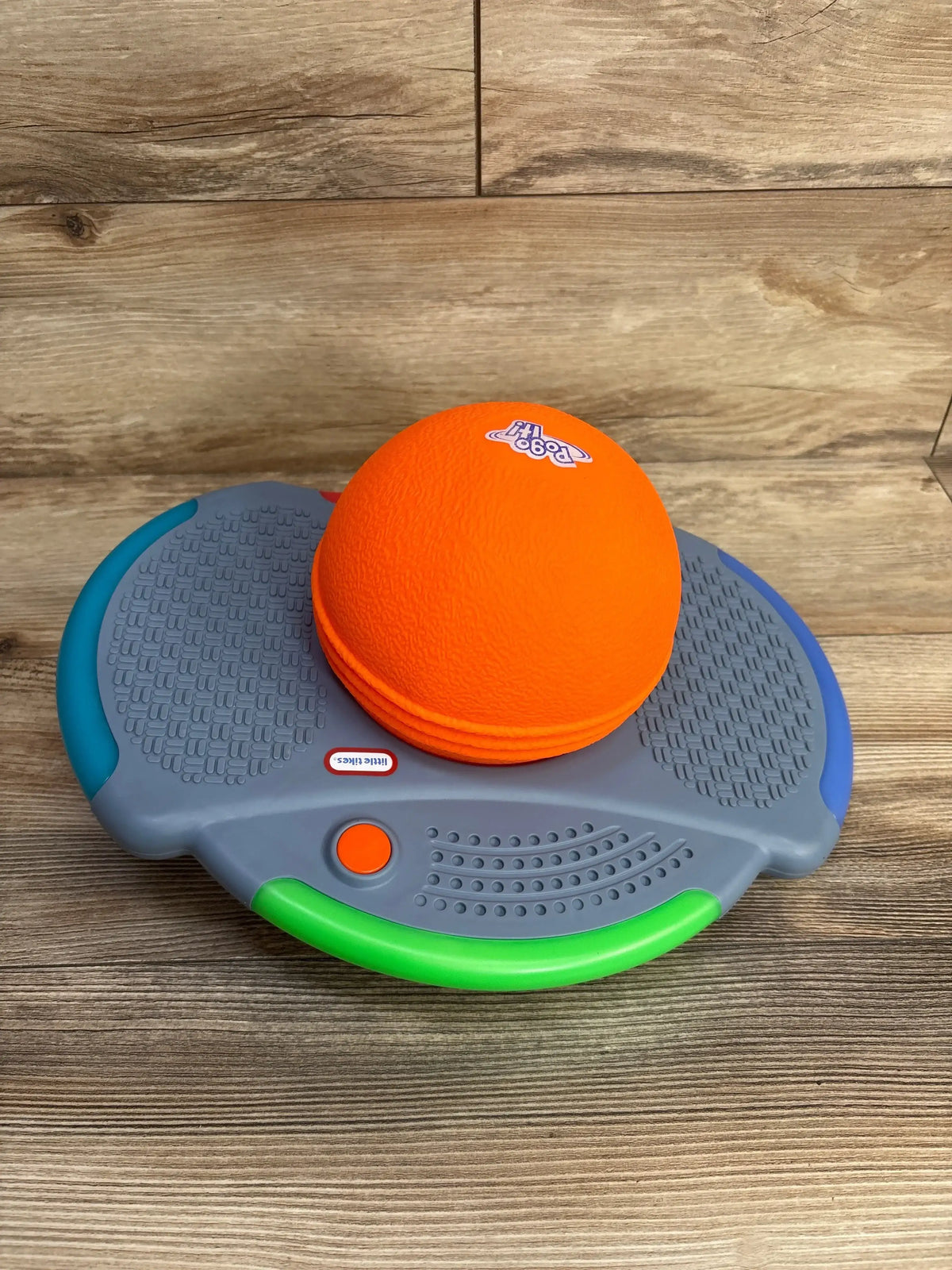 Angled top view of Little Tikes Pogo It with bright orange ball, textured gray footpads, green front handle, and blue trim on wood floor