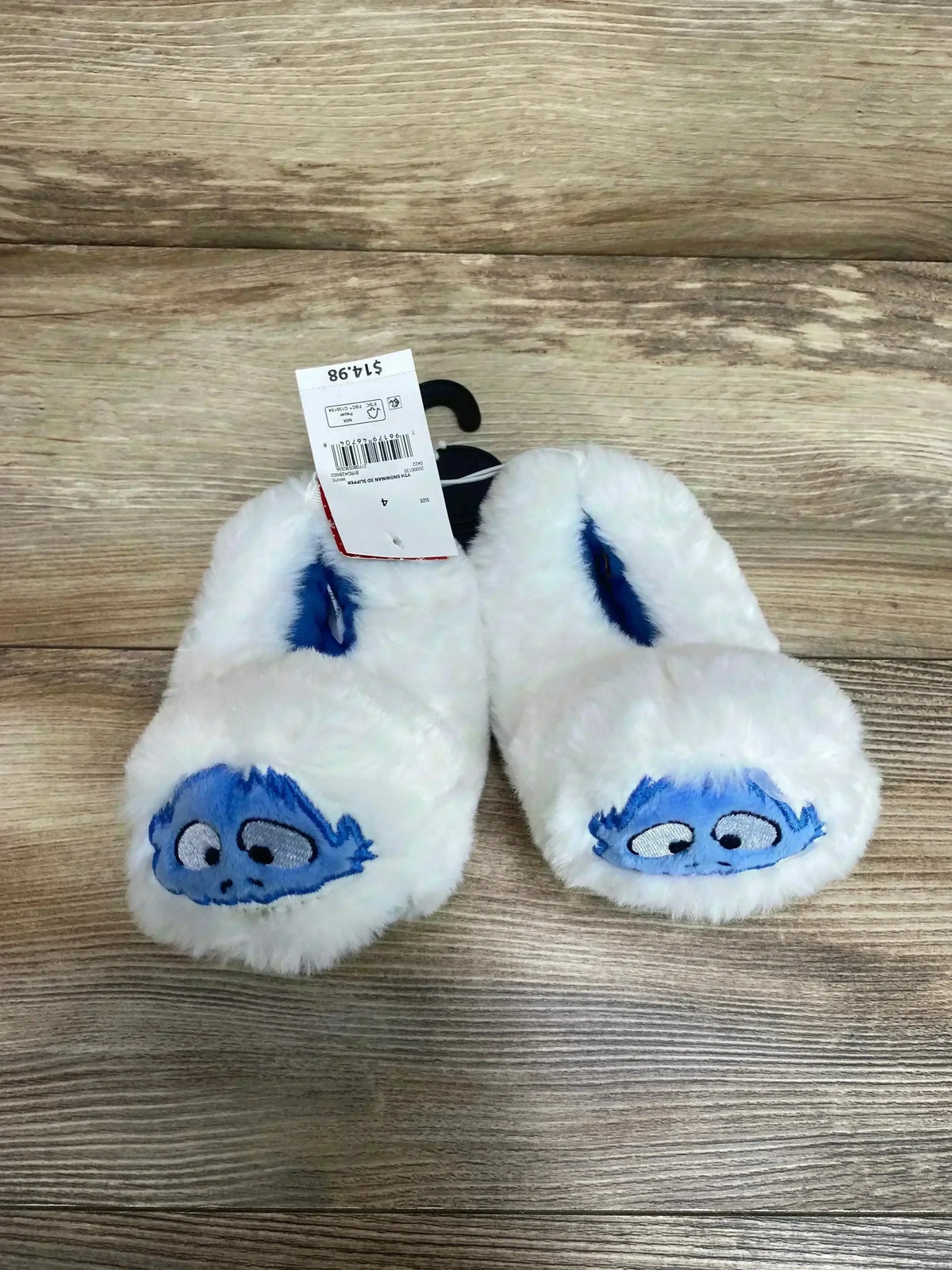 Top view of white plush toddler slippers featuring blue Abominable Snowman face embroidery on toes with wood floor background