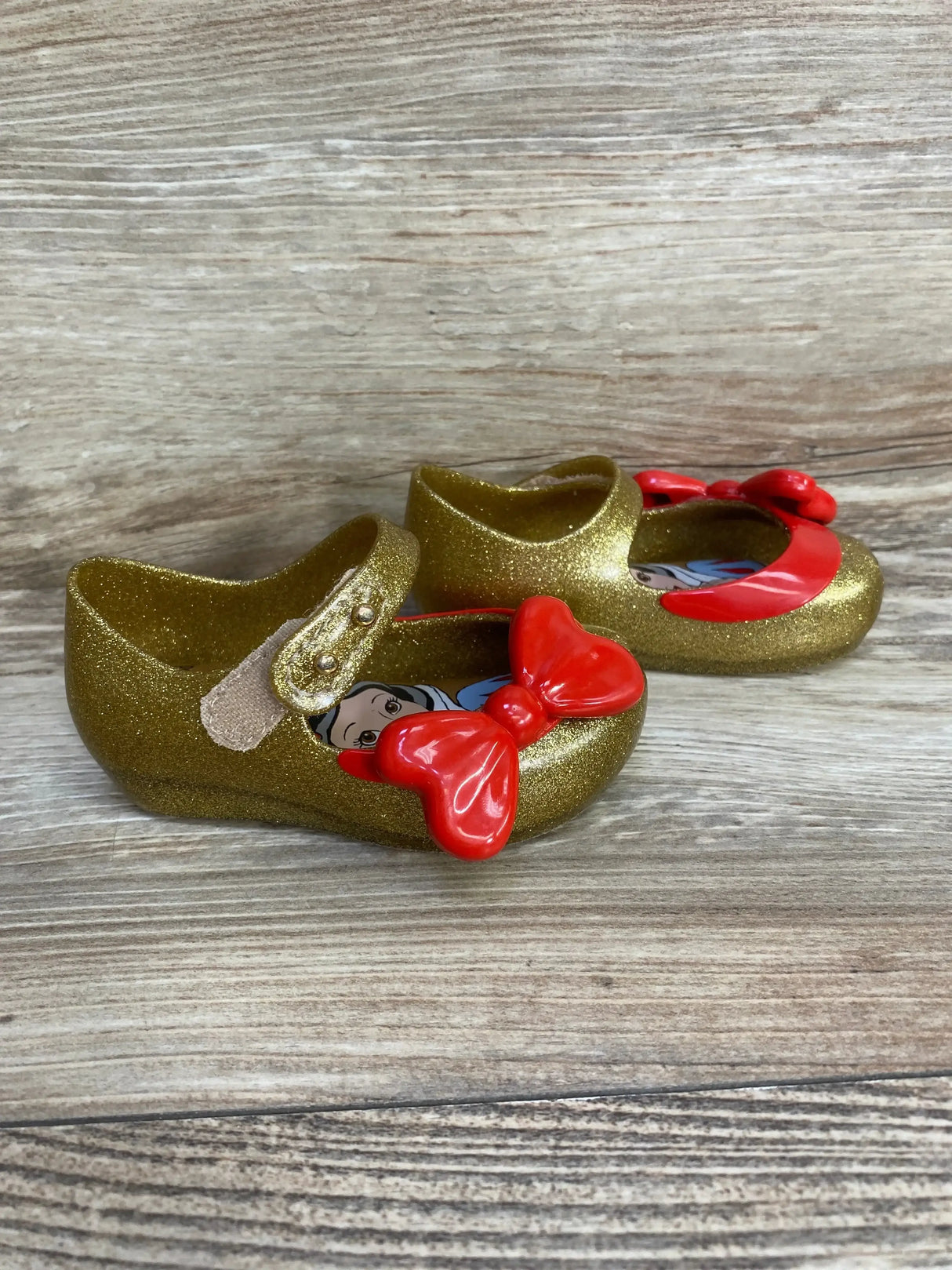 Angled front view of Mini Melissa Ultragirl toddler shoes showcasing gold glitter finish, red bow, Snow White insole on wood background