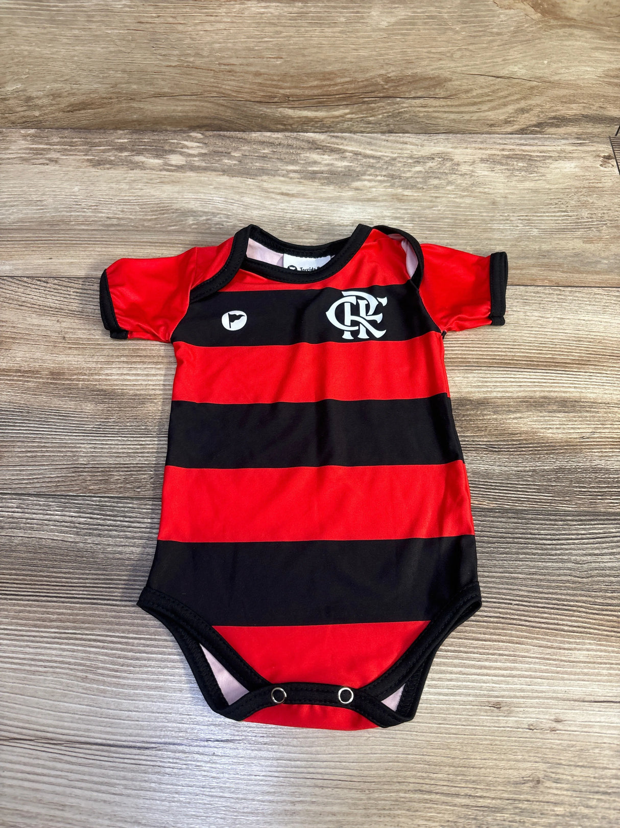 Flat lay of Torcida baby Flamengo bodysuit with red and black stripes, chest crest, short sleeves and snap crotch on wood