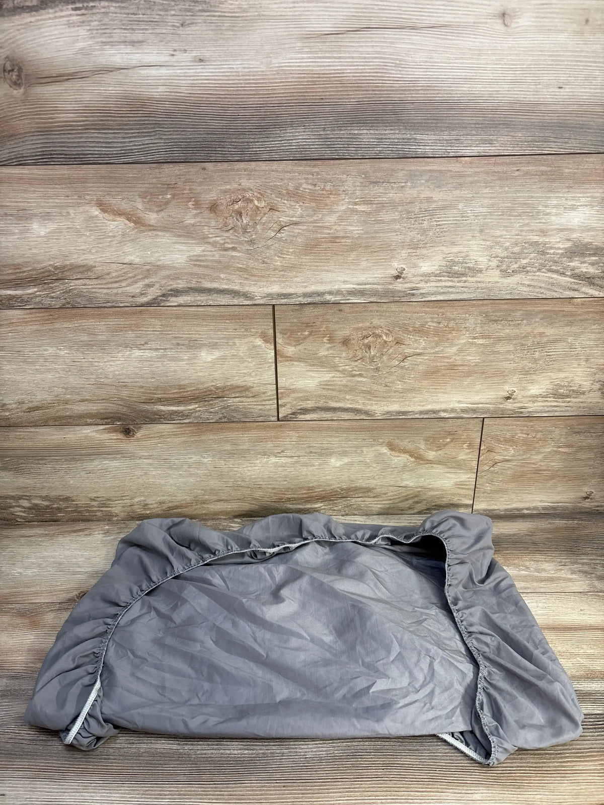 Top-down view of fitted bassinet sheet in grey showcasing elastic fitted edges and wrinkled fabric on wood plank background