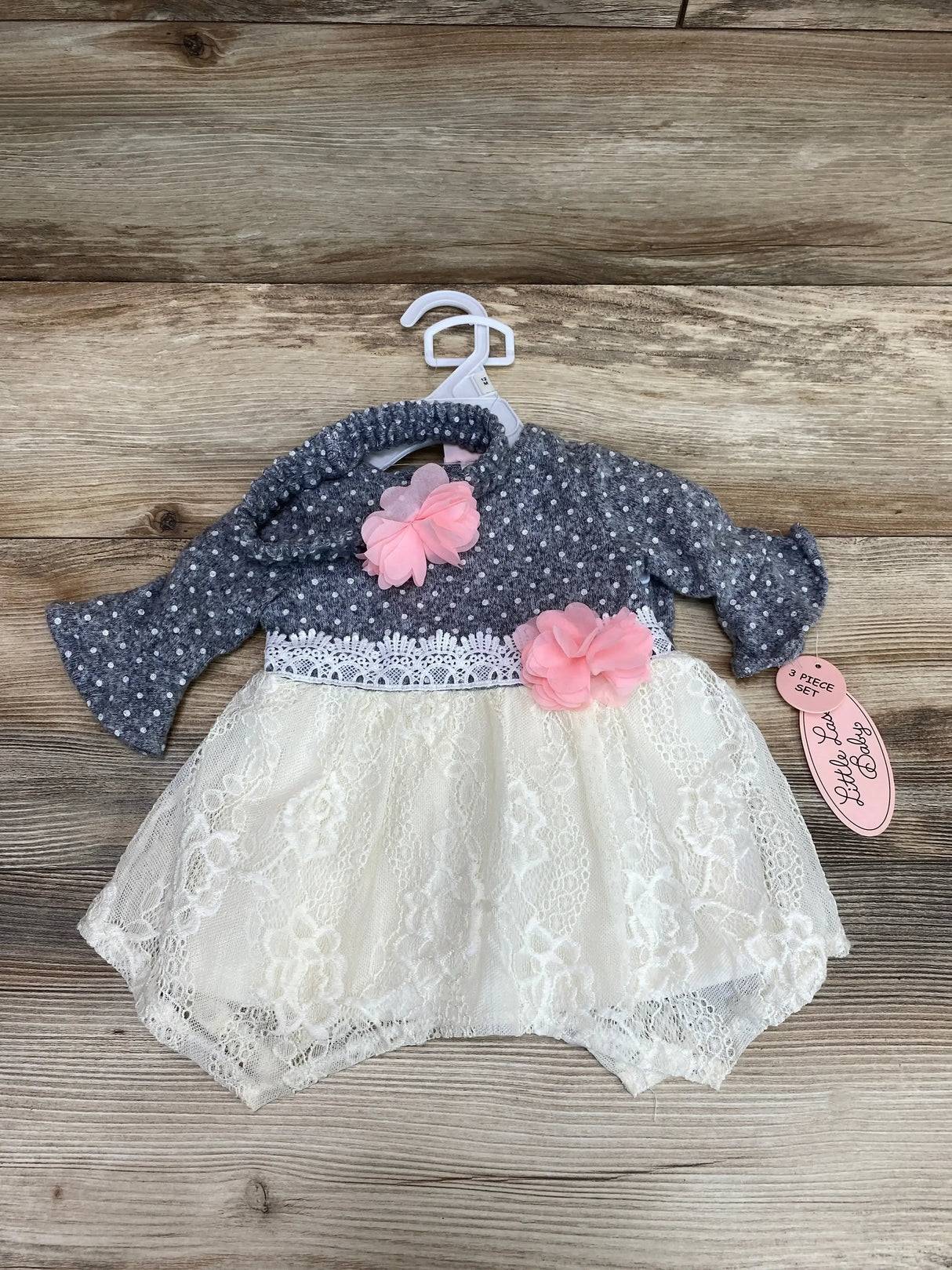 Flat lay of Little Lass 3-piece polka dot dress set showing grey long-sleeve top with white dots and pink flower accents, cream lace skirt, and matching headband on wood background