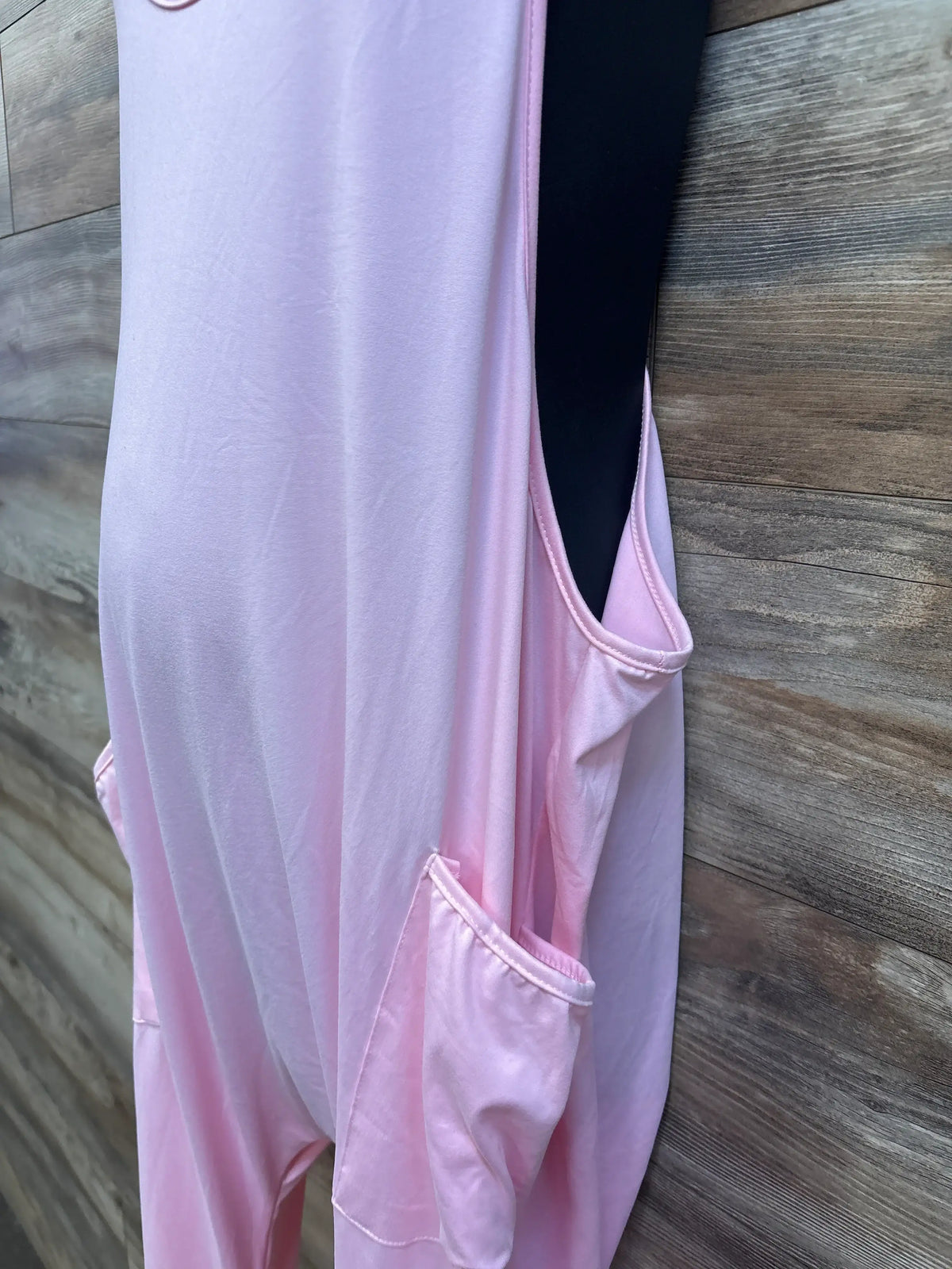 Close-up side view of pink sleeveless maternity jumpsuit showcasing a large pocket and open armhole against a wood floor