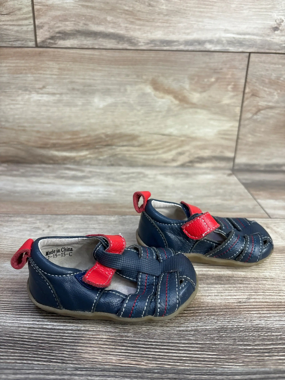 Angled front view of navy toddler fisherman sandals with red Velcro straps, woven closed toe, and flexible tan sole on wood floor
