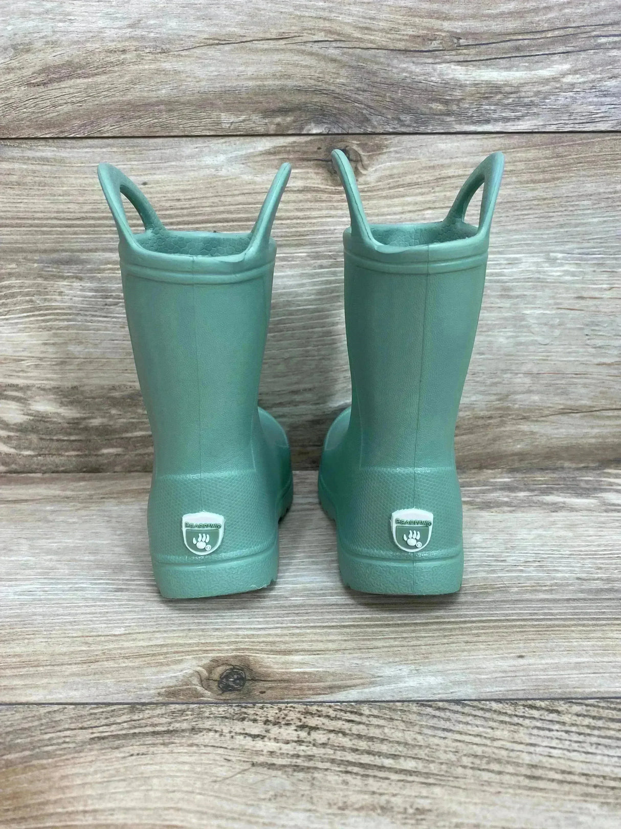 Rear view of green BEARPAW toddler EVA rain boots showing pull on handles and heel logo on wood plank background