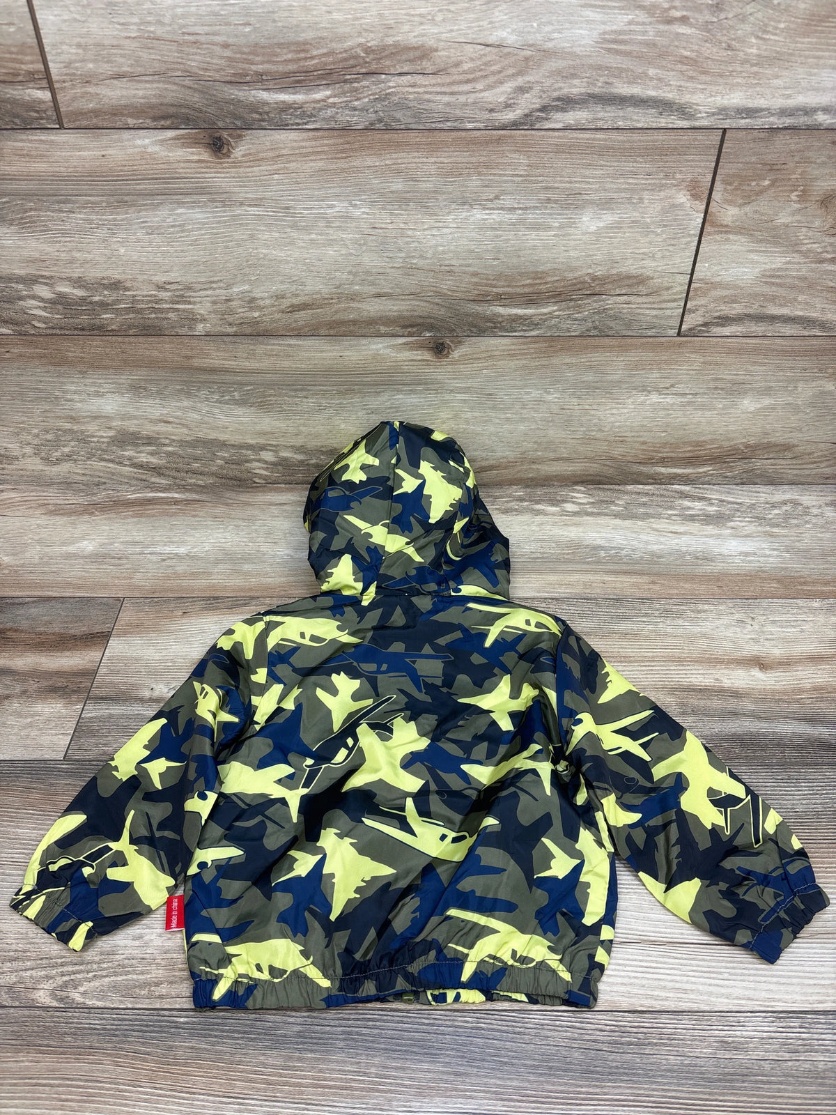 Back view of green camouflage airplane windbreaker jacket in size 18 months, with hood and elastic cuffs on wood flooring
