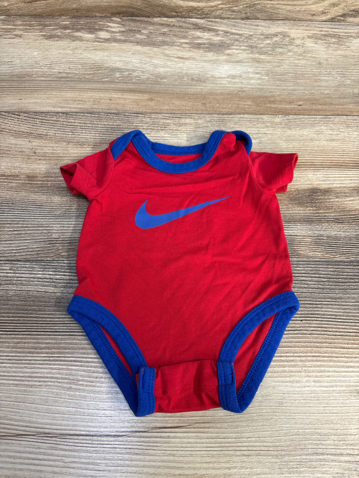 Red baby onesie with blue trim and blue Nike swoosh logo on wood background