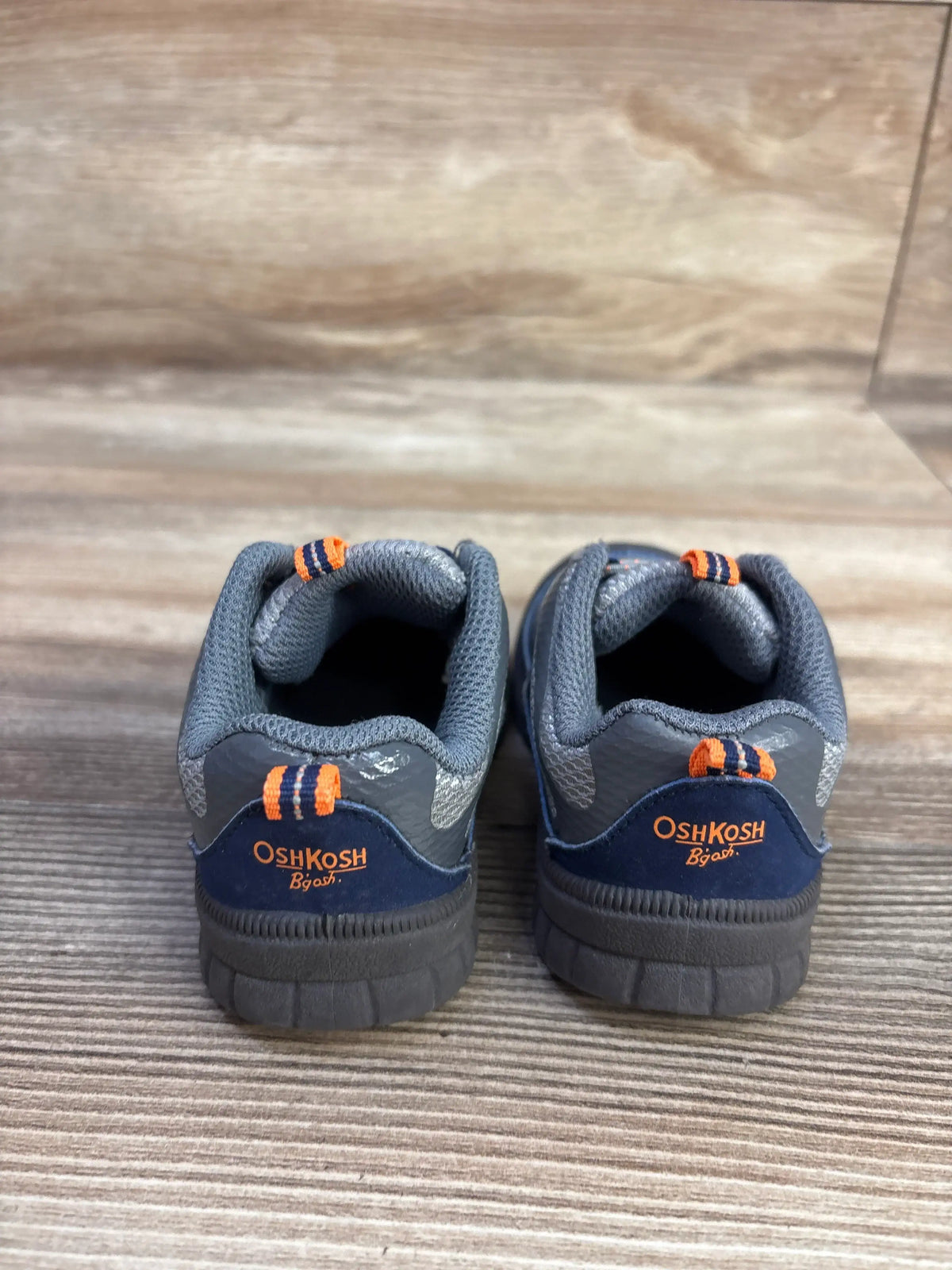 Rear view of Oshkosh Gianni bump toe sneakers showcasing padded collars and orange pull tabs on a wood-grain surface