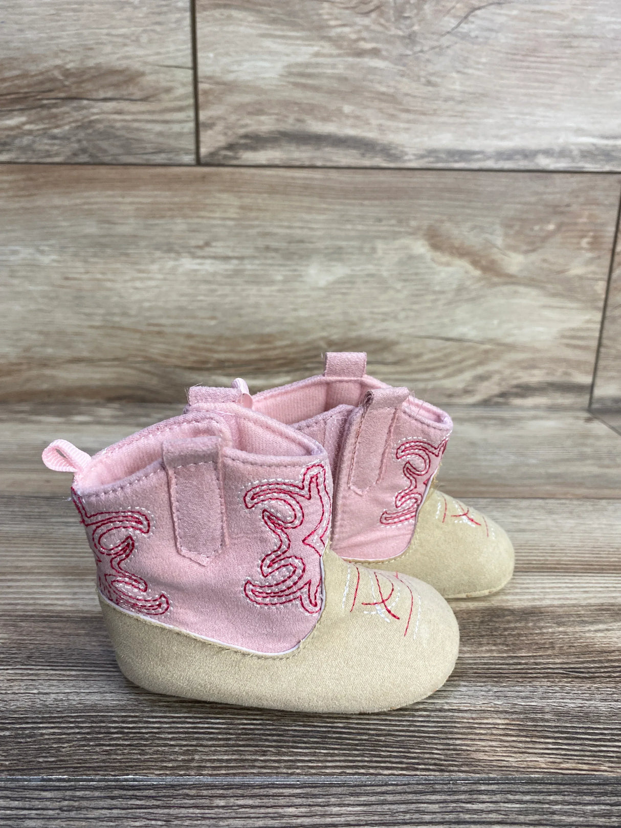 Side view of pink Stepping Stones cowgirl crib boots showcasing embroidered stitching and pull tabs on wood floor background
