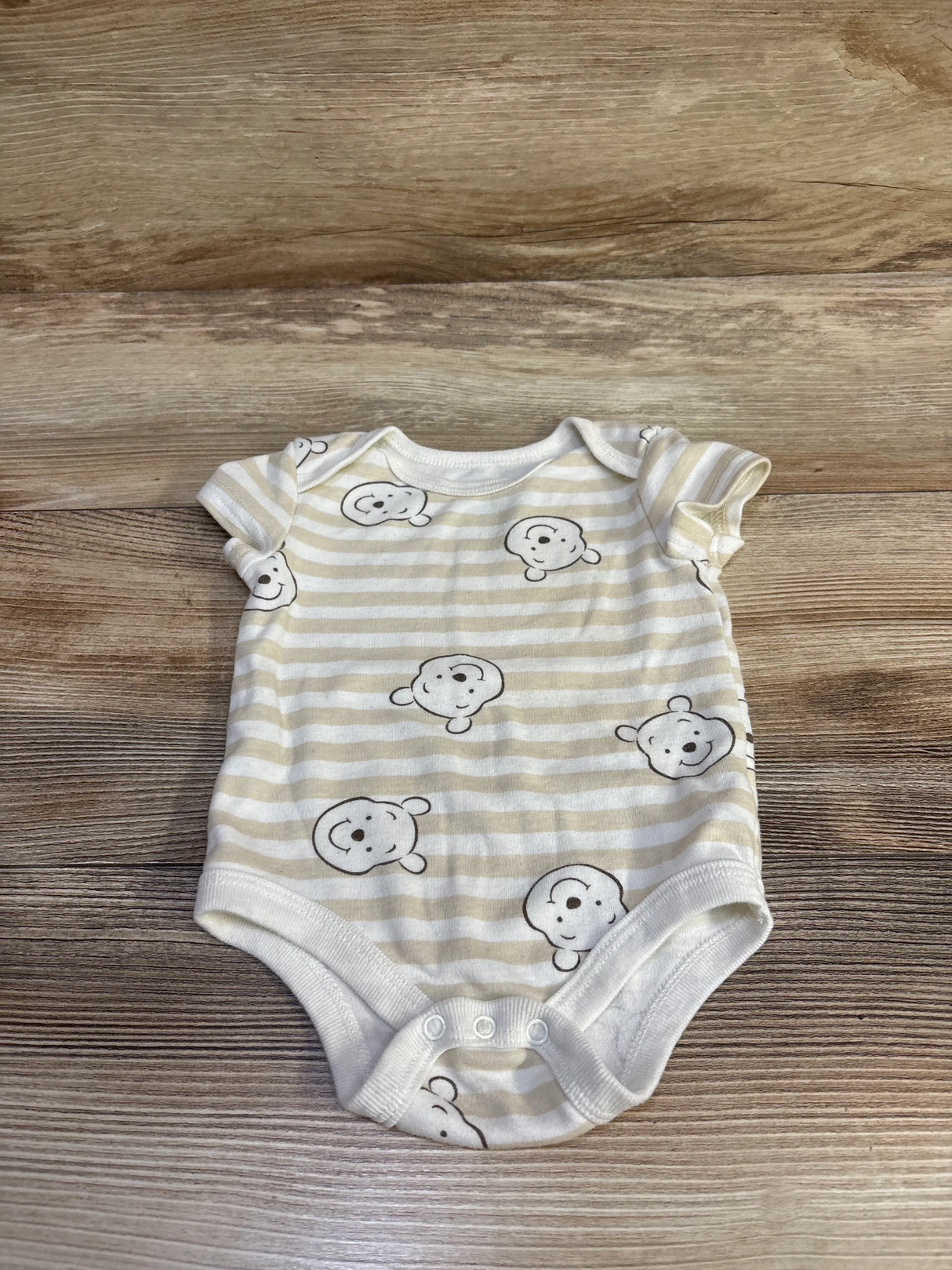 Flat lay of Disney Baby Winnie the Pooh beige striped short-sleeve bodysuit with Pooh faces and three snaps on wood