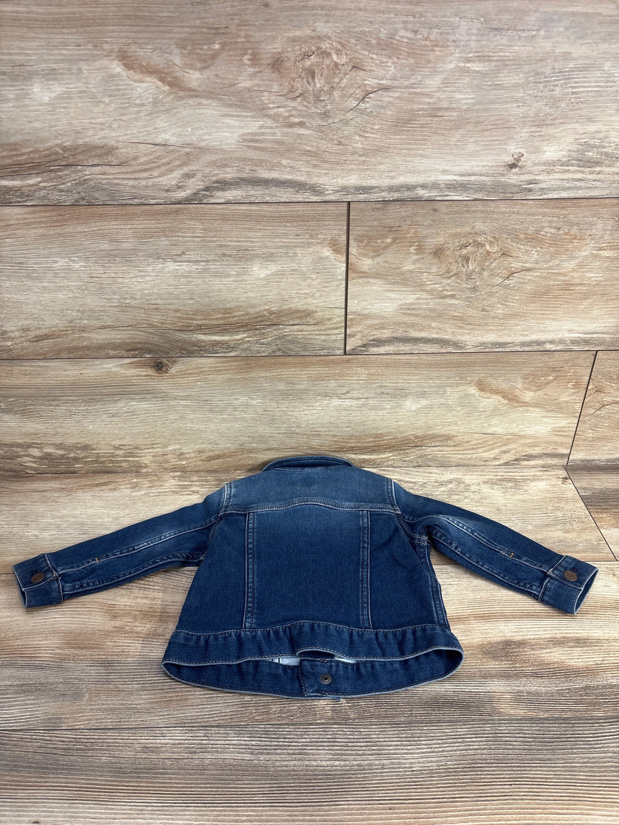 Back view of a dark blue denim jacket laid flat on wooden floor with visible stitching and button details