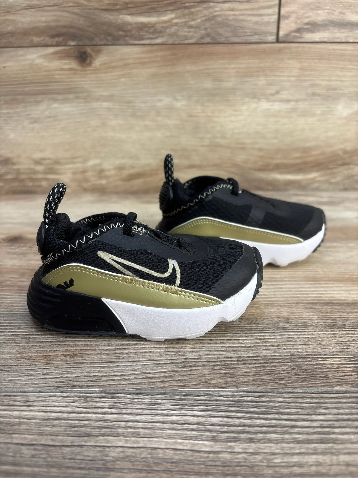 Three-quarter side view of Air Max 2090 TD toddler sneakers in black with metallic gold accent, white midsole on wood floor