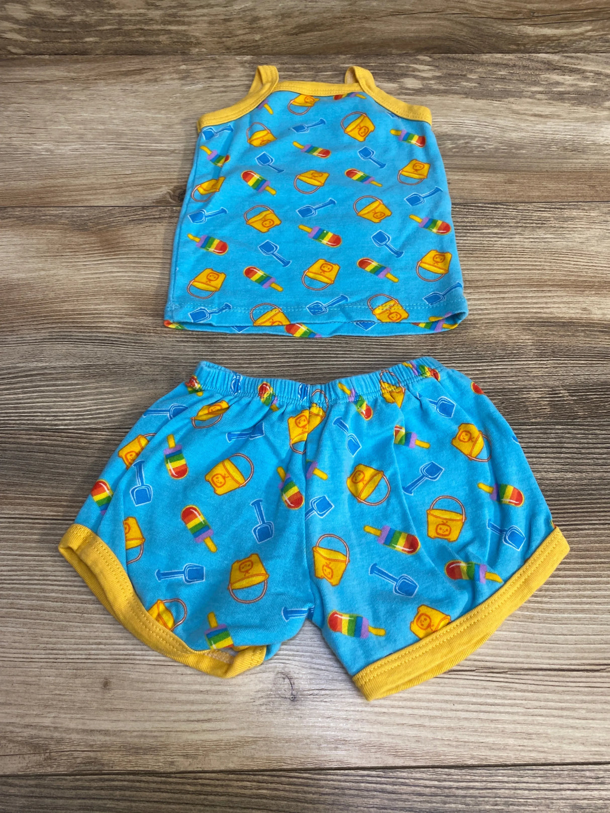 Flat lay of blue 2-piece baby tank top and shorts set with yellow trim and elastic waist, beach pail, shovel, and popsicle print on wood