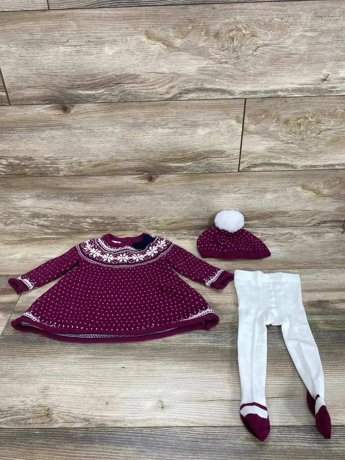 Flat lay of purple Fair Isle 3-piece knit baby dress set showing yoke pattern, pom hat, and white footed tights on wood