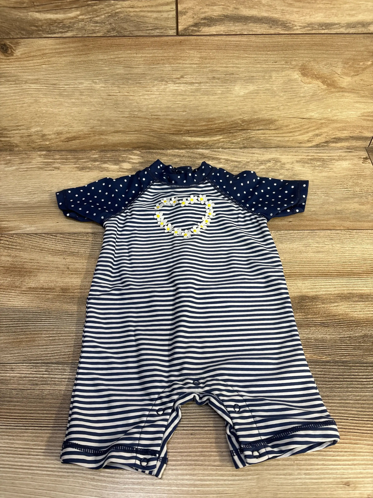 Flat lay of navy and white striped one-piece rashguard swimsuit showing navy polka-dot short sleeves, daisy-heart applique, and crotch snap closures on wood floor
