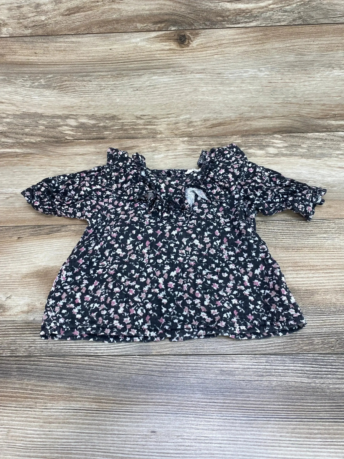 Front view of Jessica Simpson black floral ruffle top, size 12 months, showcasing ruffled sleeves on a wood background