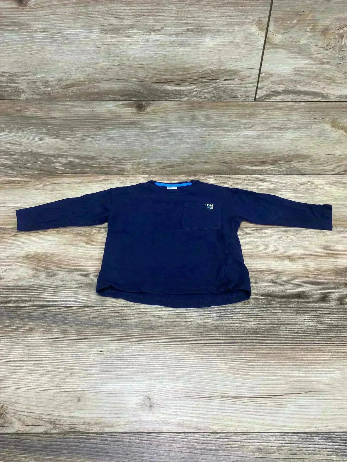 Flat lay of blue H&M long-sleeve pocket shirt (9-12 months) showcasing chest pocket on light wood floor background